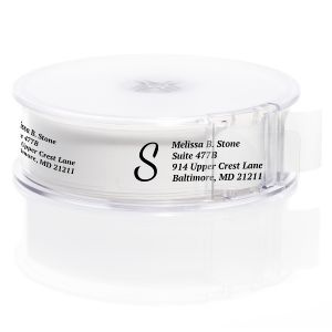 A roll of clear return address labels with a bold black letter S monogram layout, neatly enclosed inside a transparent round plastic dispenser.