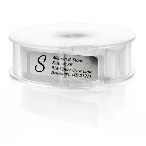 A roll of personalized silver foil return address labels with a bold black letter S monogram, housed in a clear round plastic dispenser.