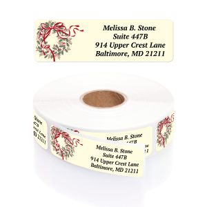 A rectangular personalized address label displayed above a large roll of matching labels, showcasing an elegant green holly and red bow wreath.