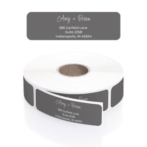 A roll of charcoal gray address labels inside a clear dispenser, showcasing white cursive names and block lettering for the address.