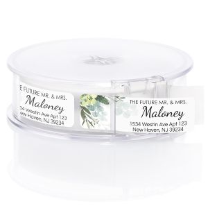 Roll of personalized wedding greenery return address labels shown in a clear round plastic dispenser for easy mailing.
