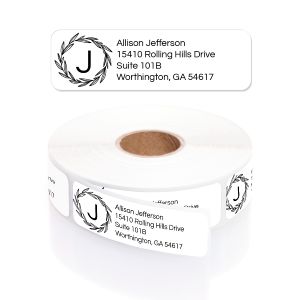 A close-up of a roll of white address labels featuring a delicate black wreath monogram on the left and clean, black text on the right.