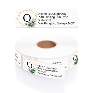 A roll of white address labels featuring a gold geometric frame and greenery wreath around a monogrammed letter 