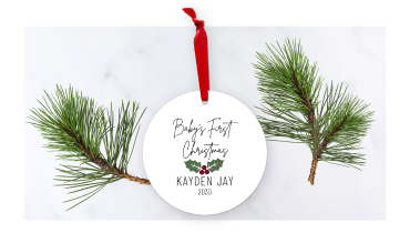 A round white ornament with a red ribbon that reads Baby's First Christmas, displayed next to festive winter pine needles.