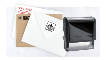 A black rectangular self-inking address stamper shown alongside envelopes stamped with crisp holiday tree graphics and custom text.